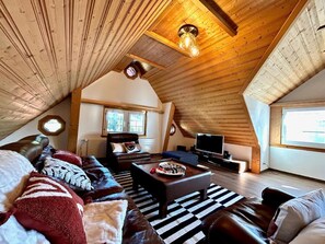 Un dormitorio acogedor en el ático, con techo inclinado, un gran ventanal, un sofá de cuero, un televisor y una chimenea.