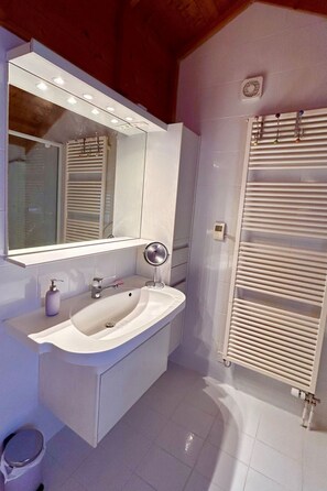 Une salle de bain moderne avec un grand miroir, un lavabo doté d’un distributeur de savon, un miroir rond posé sur un pied et un radiateur blanc.
