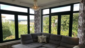 Ein Wohnzimmer mit einem grauen Ecksofa, einem runden weißen Couchtisch und großen Fenstern mit Blick auf Grünflächen und Gebäude.