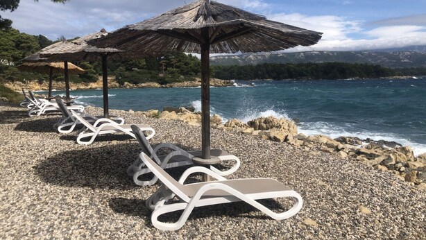 Ein Strand mit Liegestühlen und Strohdächern, mit Blick auf eine felsige Küste und das Meer.