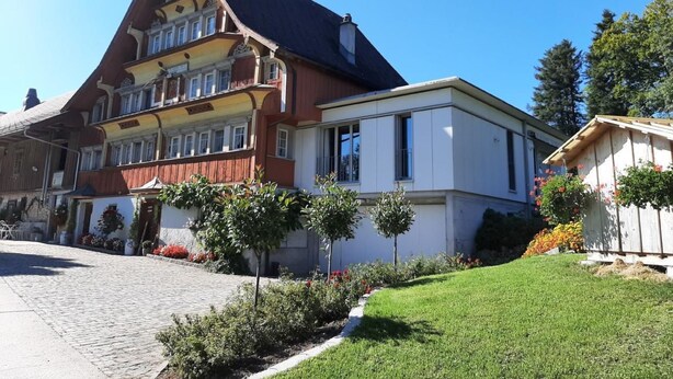 A traditional multi-story building with a steeply pitched roof, a balcony on the upper floor, and a well-maintained garden with small trees and flowers. - auf dem Bauernhof by Interhome (Lütisburg Station)