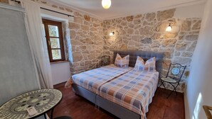 A bedroom with a stone wall, a bed with a plaid bedspread, a round table with a chair, and a small table with a decorative item. - Tolles Ferienhaus in Supetarska Draga mit Garten by Interhome (Rab/Supetarska Draga)