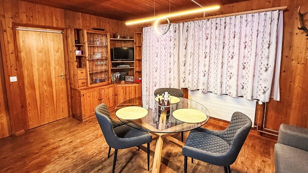 Een kamer met houten lambrisering, een rond glazen tafeltje, twee stoelen, een kast met planken en een groot raam met gordijnen.