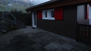 Een gebouw met een bruine buitenkant, een witte deur en rode luiken. Een grindpad leidt naar een reeks trappen.
