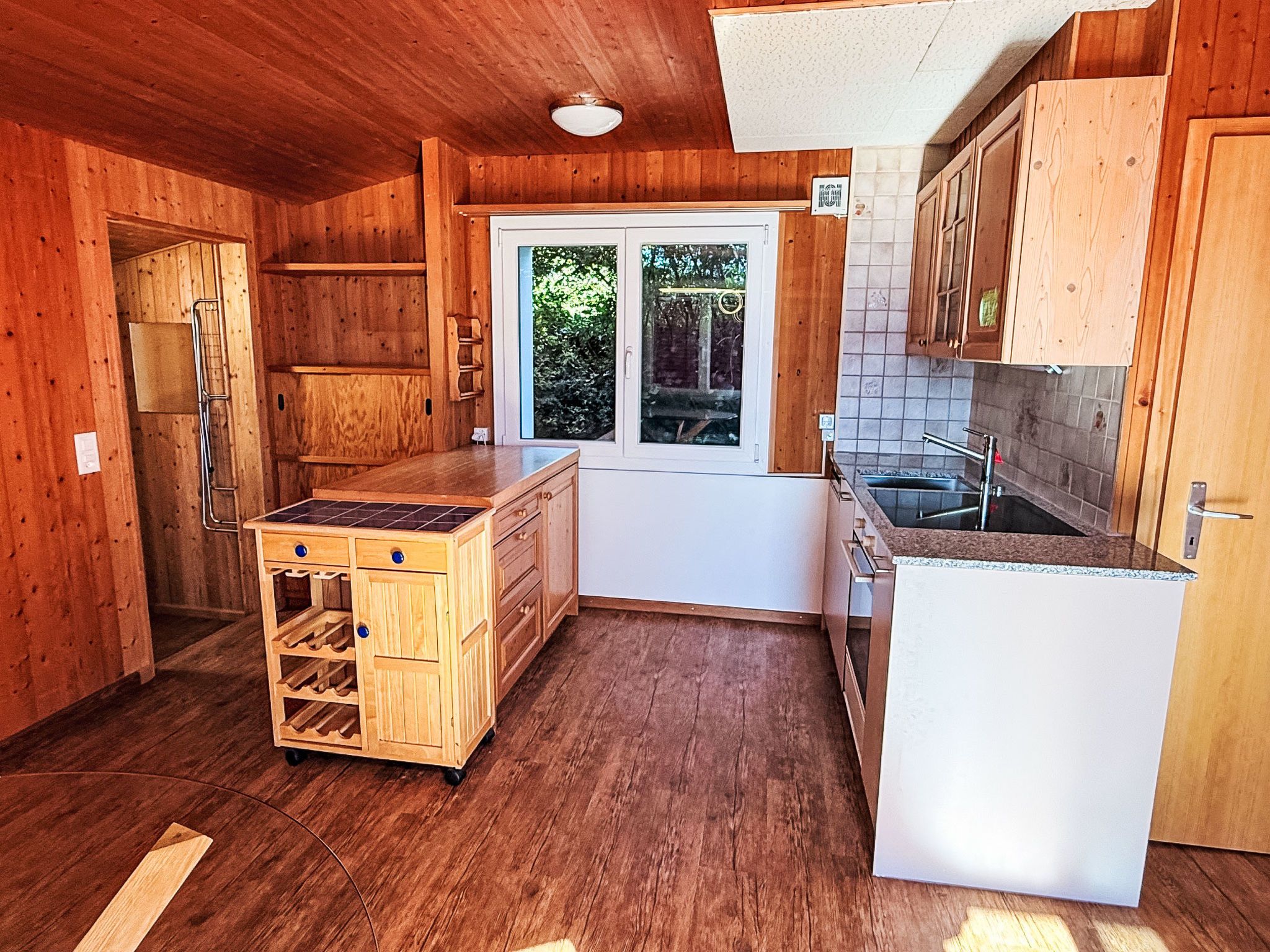 Cocina compacta con muebles de madera, pileta integrada y ventana con vista a follaje.