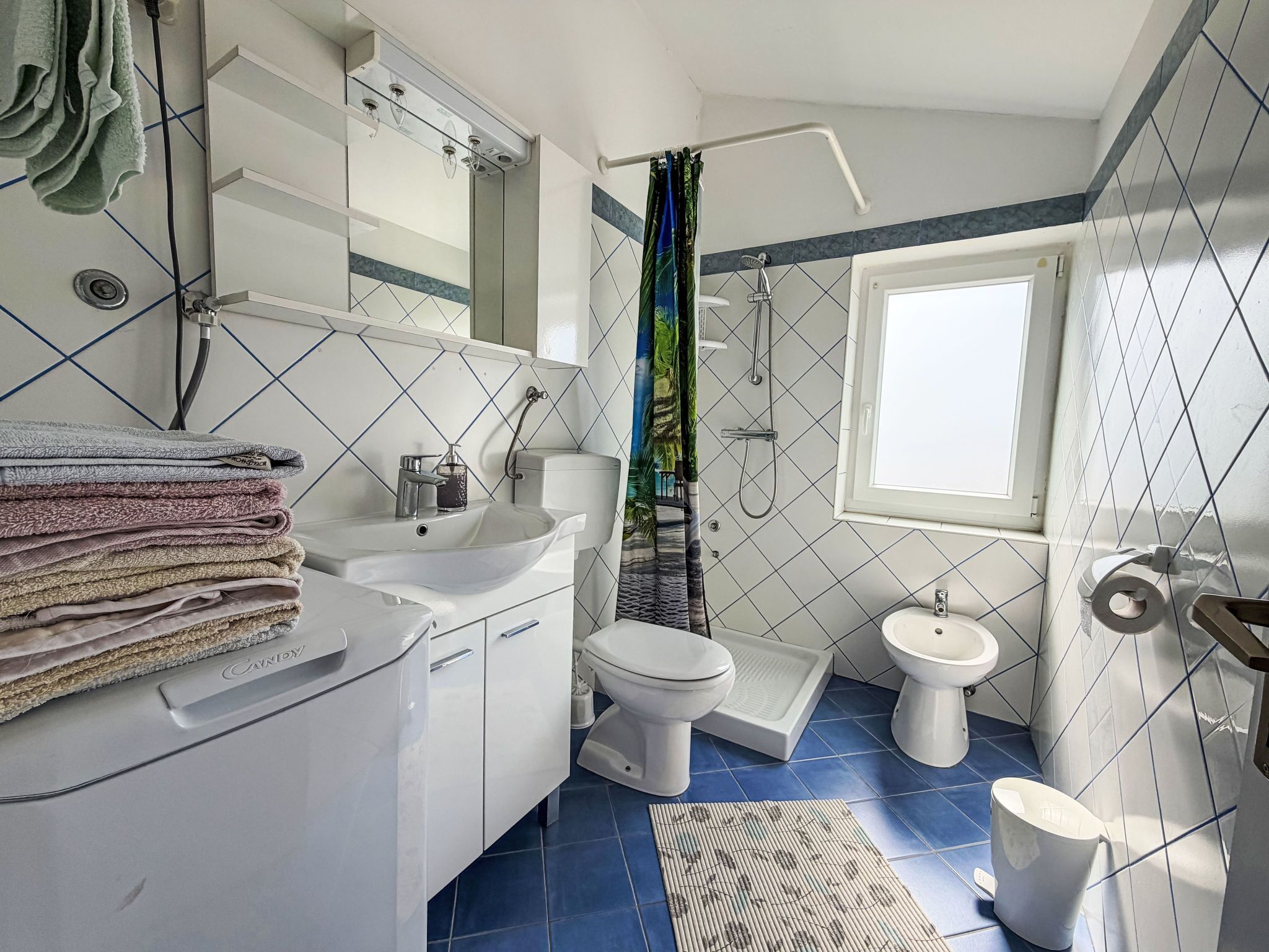 Une salle de bain équipée d’un lavabo, de toilettes et d’une douche. On y trouve un porte-serviettes avec des serviettes, une fenêtre et un miroir.