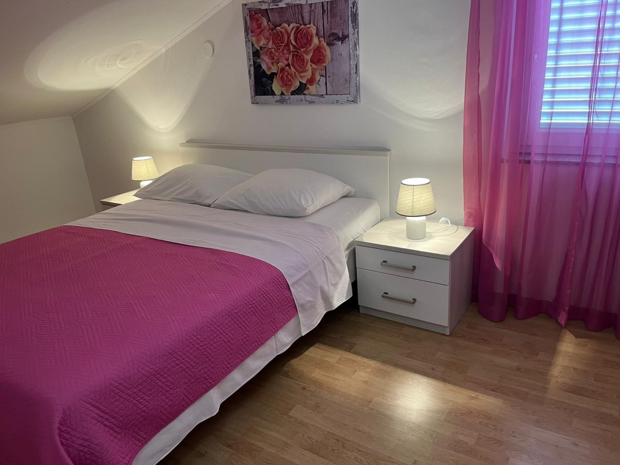 Un dormitorio con una cama, dos mesitas de noche con lámparas, una cortina rosa y una obra de arte floral en la pared.