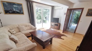 Ein Wohnzimmer mit einem beigefarbenen Sofa, einem Holztisch und einem Ledermatte. Ein Fernseher steht auf einem Holzgestell, und eine Tür führt nach draußen.