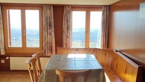 Un comedor con mesa y bancos, ventanas amplias con cortinas y vistas al paisaje.