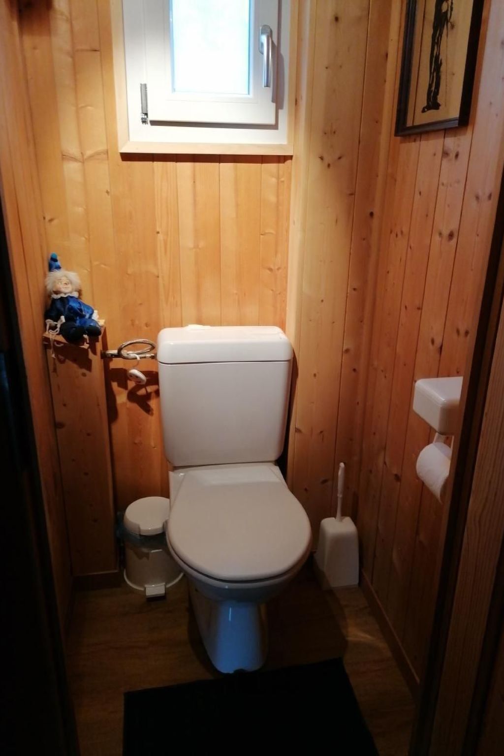 A small, wooden-paneled bathroom with a toilet, a small trash bin, and a window above the toilet.