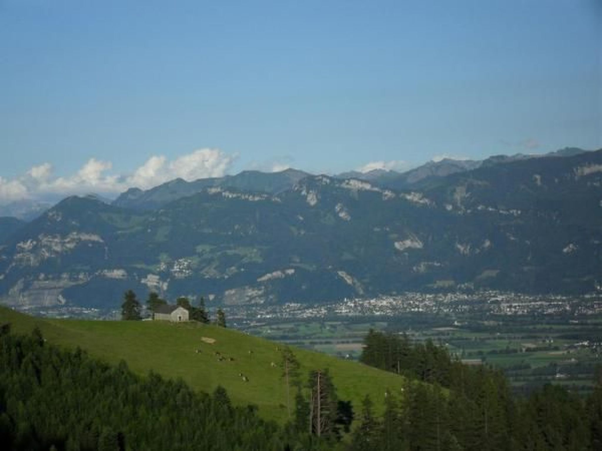 Een berglandschap met een klein huis op een heuvel, omringd door groen en bergen in de verte.