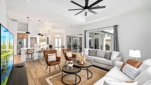 A modern living room with a sofa, coffee table, and chairs. There is a TV, a ceiling fan, and a kitchen in the background. - South Cape Luxury | Putt Green, HTD POOL,& Game RM (Cape Coral)