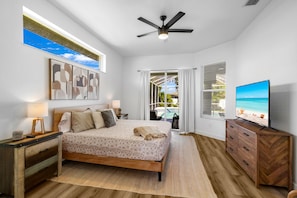 A bedroom with a bed, bedside tables, a ceiling fan, a TV, and a view of a pool and beach. - South Cape Luxury | Putt Green, HTD POOL,& Game RM (Cape Coral)