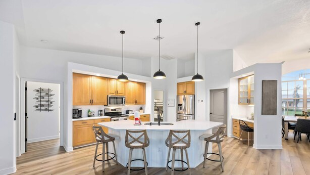 A modern kitchen with wooden cabinets, a central island with bar stools, pendant lighting, and a view of the outdoors. - South Cape Luxury | Putt Green, HTD POOL,& Game RM (Cape Coral)