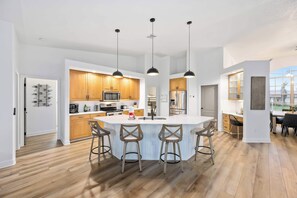 A modern kitchen with wooden cabinets, a central island with bar stools, pendant lighting, and a view of the outdoors. - South Cape Luxury | Putt Green, HTD POOL,& Game RM (Cape Coral)