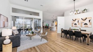 A spacious living area with a dining table, chairs, and a sofa. There is a kitchen area in the background. - South Cape Luxury | Putt Green, HTD POOL,& Game RM (Cape Coral)