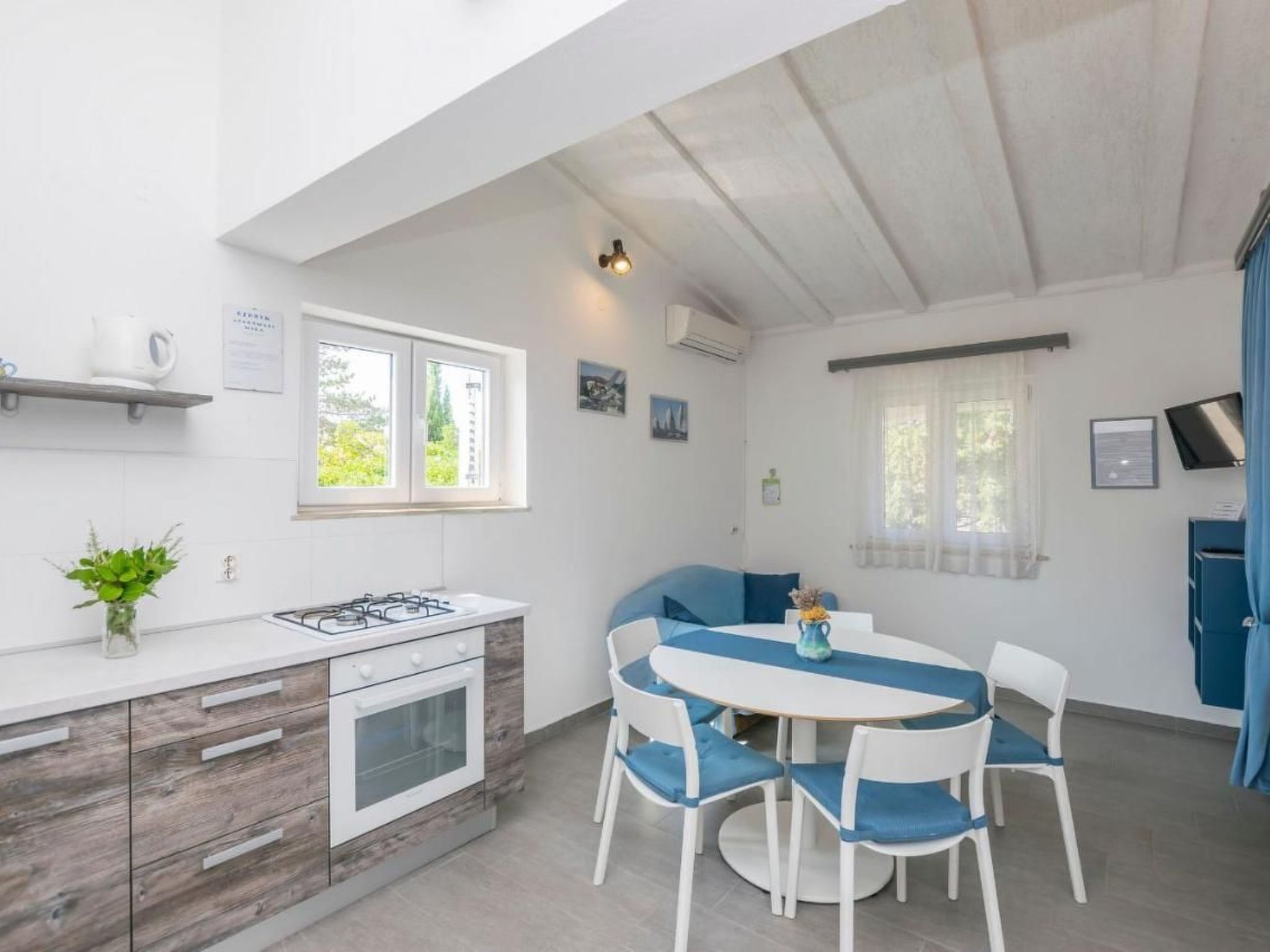 Eine kompakte Küche mit Gasherd, Backofen und kleinem Fenster. Es gibt einen Essbereich mit rundem Tisch und Stühlen, sowie ein blaues Sofa.