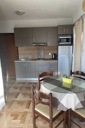 Een compacte keuken met grijze kastjes, een magnetron en een koelkast. Er staat een rond glazen tafel met stoelen en een vaas met gele bloemen.