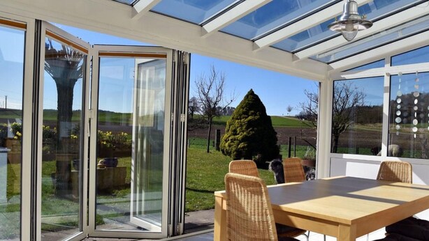 Une véranda avec un toit en verre, une table en bois, des chaises en osier et une vue sur un jardin.