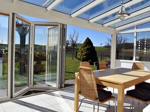 Ein Wintergarten mit Glasveranda, einem Holztisch, Korbstühlen und Blick auf einen Garten.
