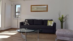 Un salon moderne avec un canapé gris foncé, une table basse en verre, une petite table ronde, un fauteuil violet et un vase avec des fleurs vertes.