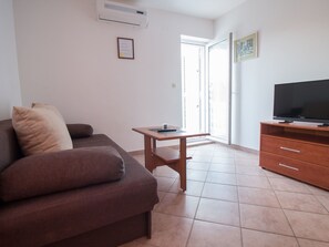 Sala de estar com sofá marrom, mesa de madeira pequena, TV em suporte de madeira e porta de vidro deslizante que dá para uma varanda.