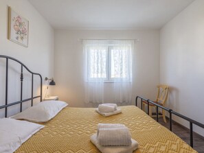 Ein Schlafzimmer mit einem Bett, Kissen, einem Nachttisch mit Lampe, einem Fenster mit durchsichtigen Vorhängen und einem Sessel.