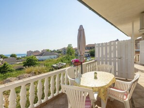 Eine überdachte Terrasse im Freien mit einem runden Tisch und weißen Stühlen, mit Blick auf ein Wohngebiet und das Meer.