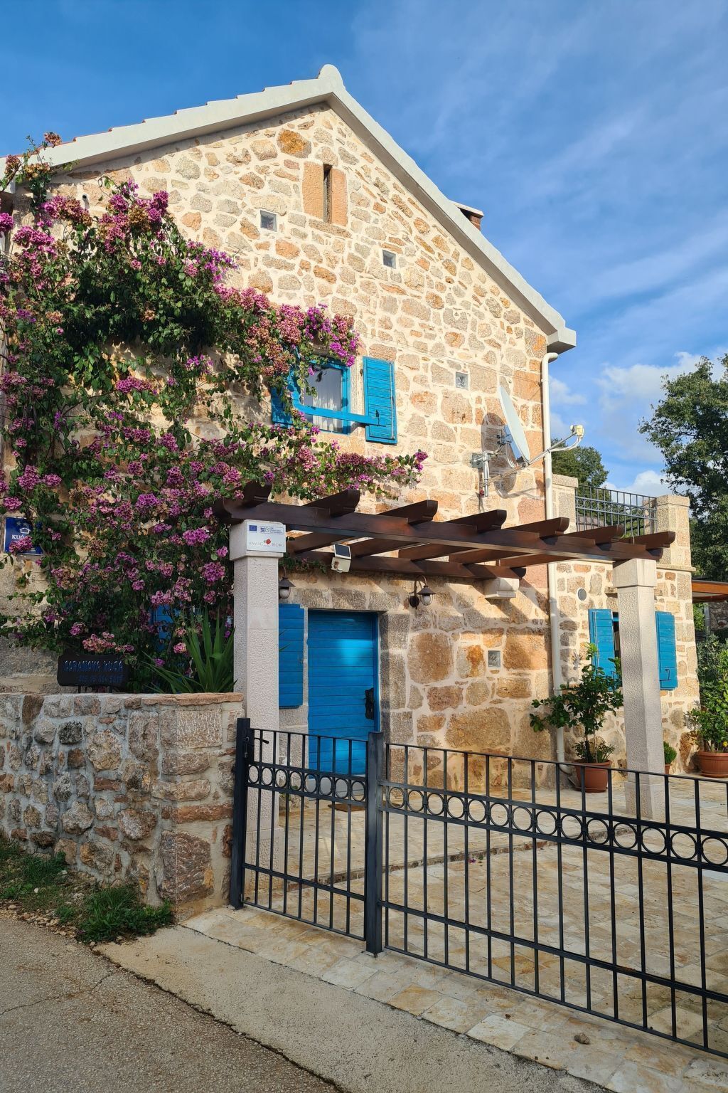 A stone building with a blue door and shutters, surrounded by a metal fence and potted plants.