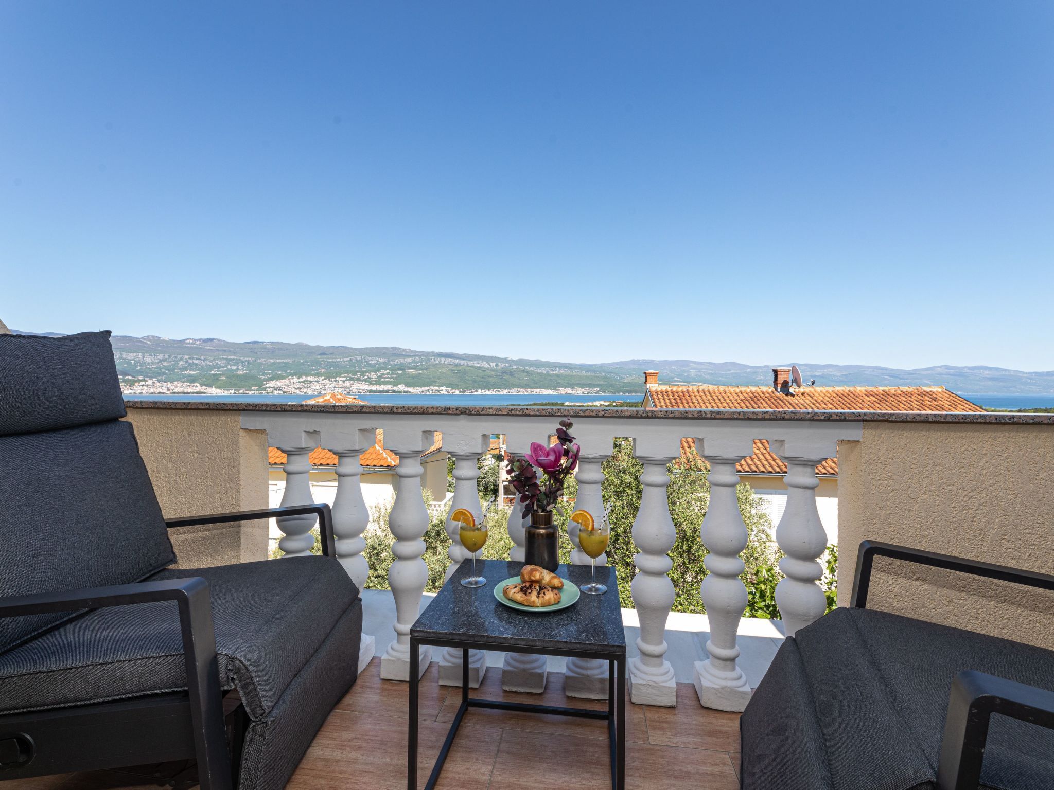 A terrace with a table and chairs, a view of the sea, and a mountainous landscape in the distance.