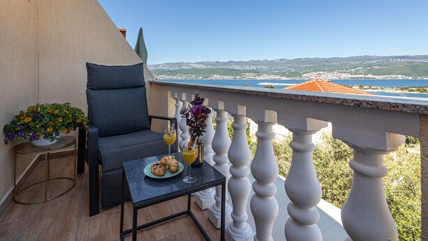 Un balcone con vista su una cittadina costiera, un tavolo con un cornetto e succo di frutta, e una sedia.