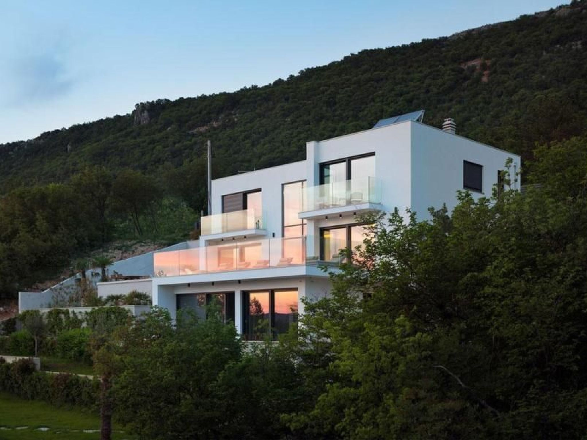 A modern two-story house with a balcony, surrounded by greenery and hills.