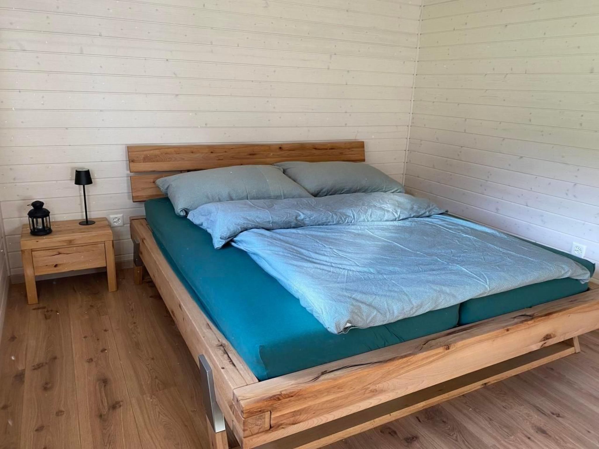 A wooden bed with a green mattress and two pillows, a small wooden nightstand with a lamp, and a white brick wall.