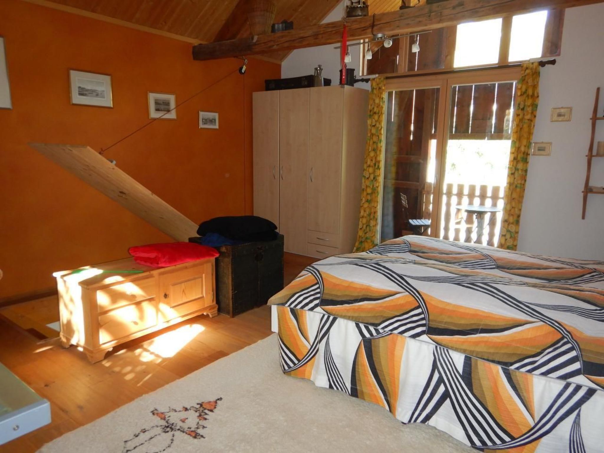 A bedroom with a bed, a wooden wardrobe, a small table with a red pillow, and a window with curtains.