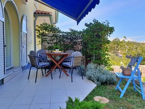 A patio with a table and chairs, a blue umbrella, and a garden with plants and a fence. - Apartment Savic by Interhome (Krk/Šilo)