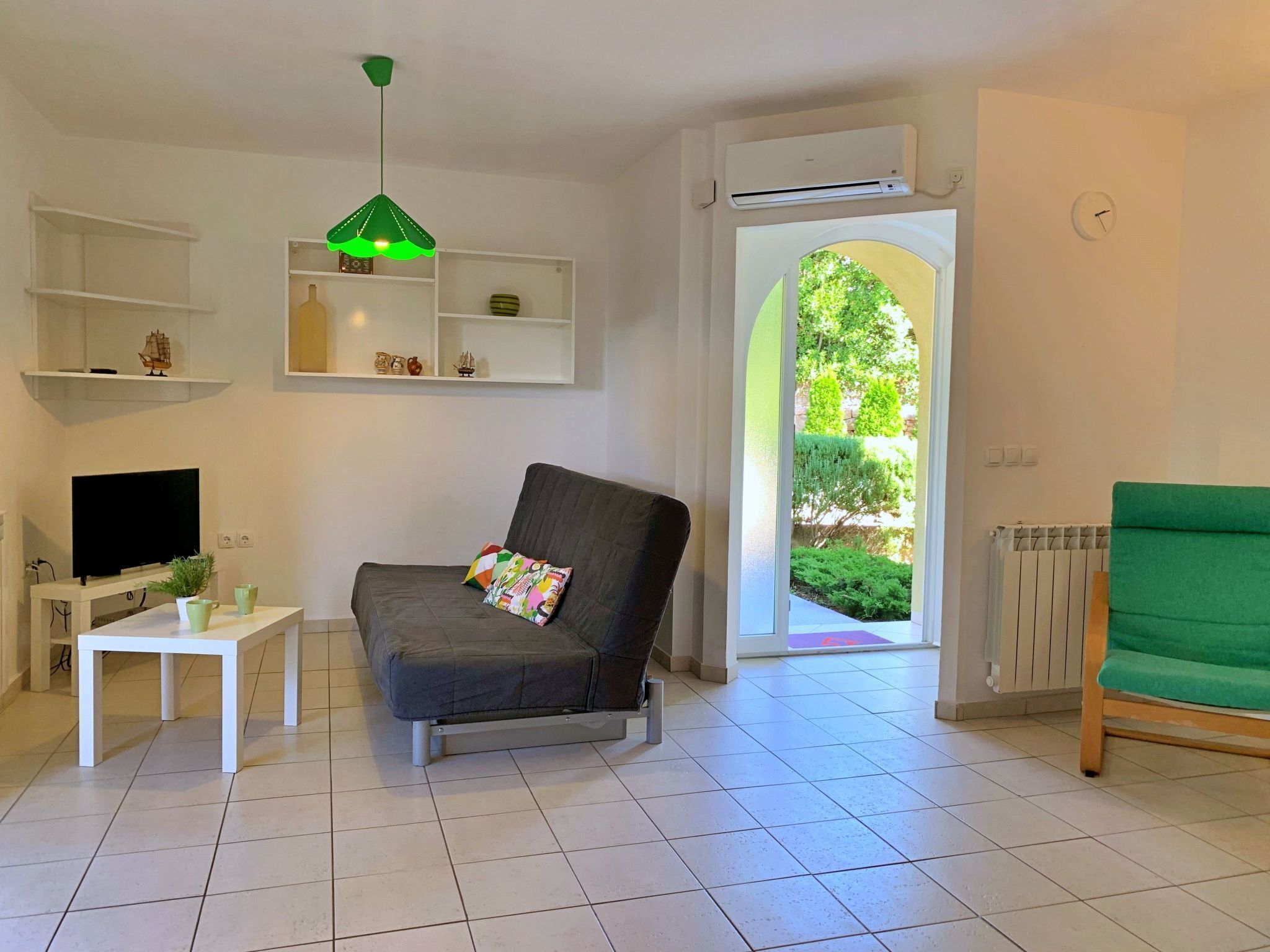 En stue med sofa, fjernsyn, et lille bord med en plante og en grøn lænestol. Der er et vindue med udsigt til grøn natur.
