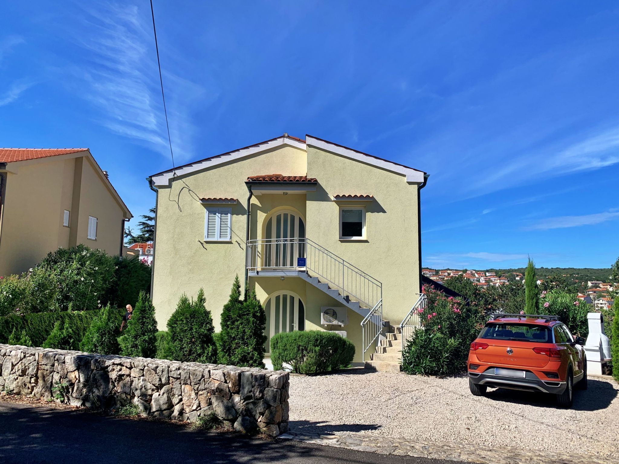 Un bâtiment à deux étages doté d’un balcon, un mur de pierre en premier plan et une voiture rouge garée devant.