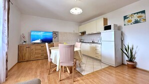 Une cuisine moderne avec des armoires blanches, un four à micro-ondes encastré et un réfrigérateur. On y trouve également un coin repas avec une table et des chaises, ainsi qu’un téléviseur diffusant une vue panoramique.