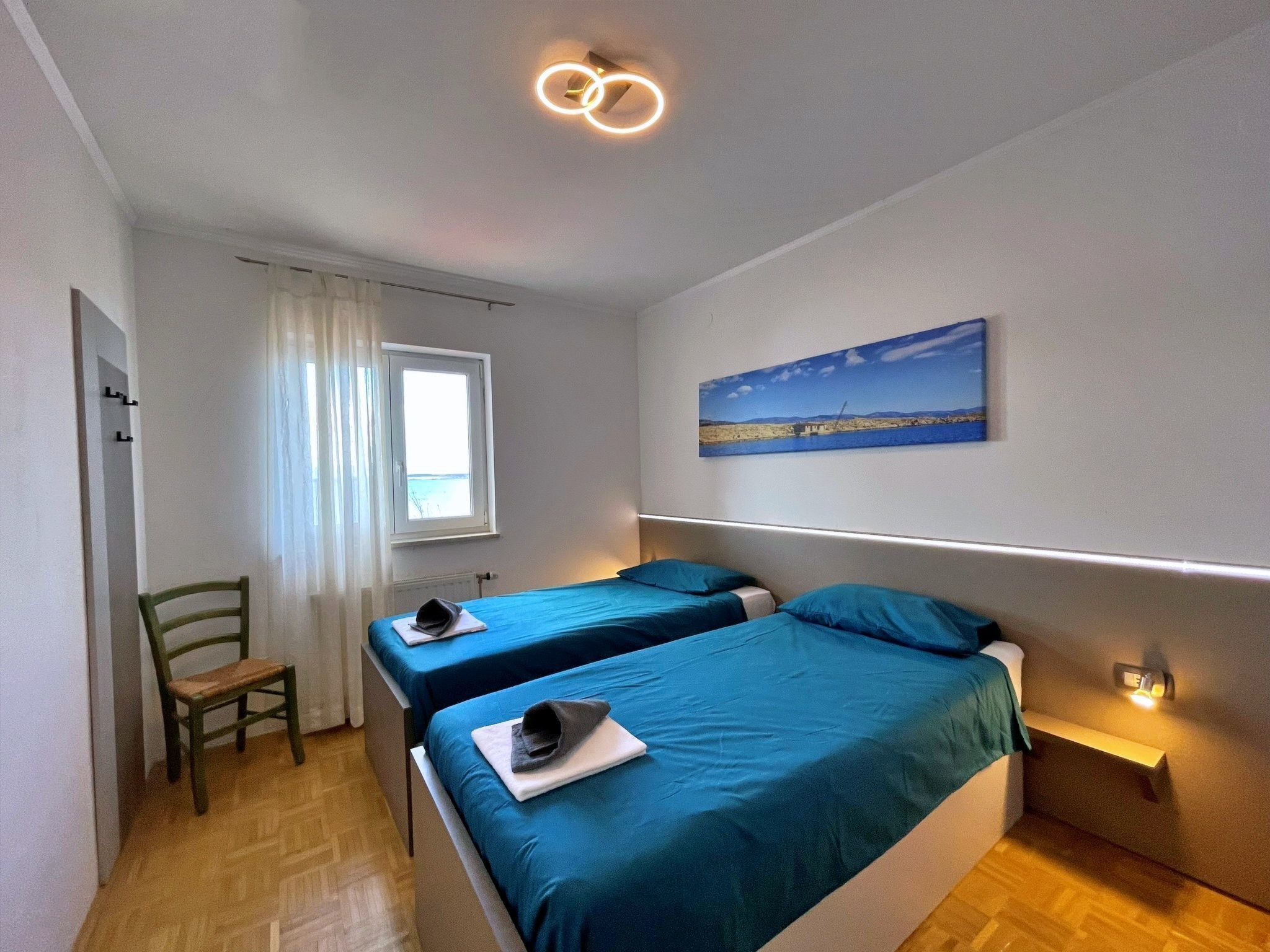 A bedroom with two beds, a chair, a window, a painting, and a ceiling light fixture.