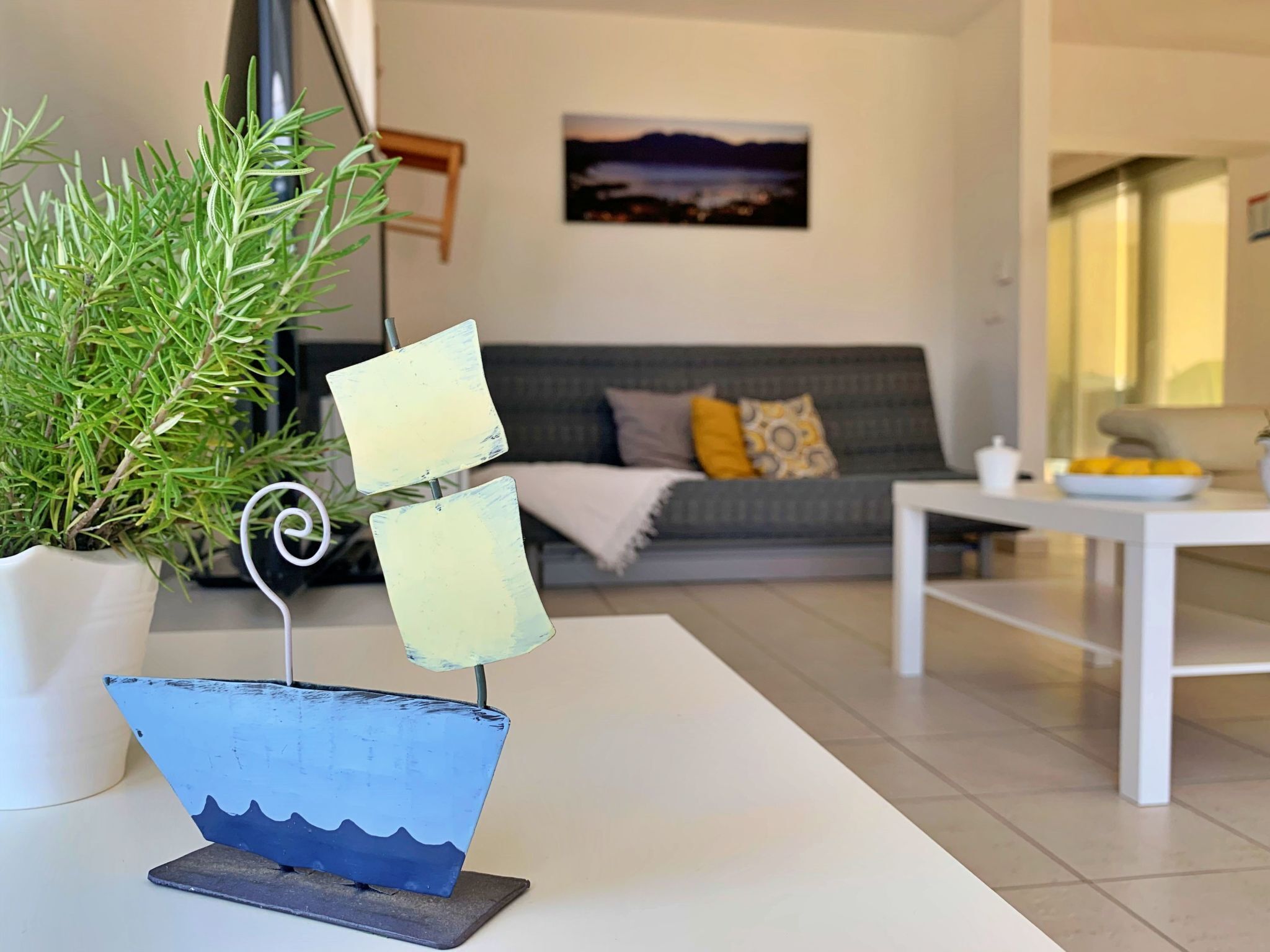A modern living room with a white table, a potted plant, a grey sofa with cushions, and a painting on the wall.
