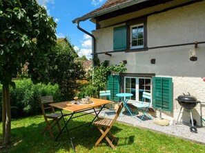 A patio with a wooden table and chairs, a barbecue grill, and a garden with various plants and a tree.