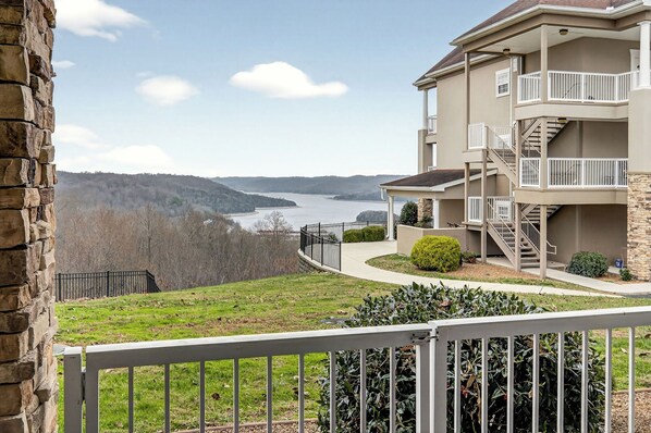 A multi-story building with a stone facade, a balcony with a white railing, and a view of a lake surrounded by hills.