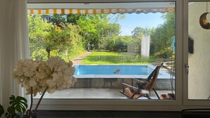 Udsigt gennem et skydedørsvindue til en baghave med swimmingpool, en stol og en potteplante.