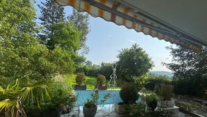 Udsigt fra en overdækket balkon ned til en swimmingpool, omgivet af krukker med planter og grønt.