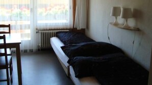 A single bed with a dark blanket, a small table, and a chair in a room with a window and a radiator. - Hof Eggweid by Interhome (Aesch ZH)