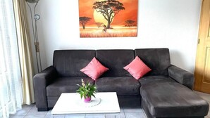 Un salon avec un canapé d’angle gris foncé, deux coussins rouges, une table basse blanche et une plante en pot. Un grand tableau représentant une scène de savane est accroché au mur.