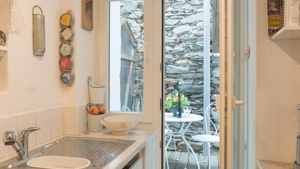 Une cuisine équipée d’un évier, d’une fenêtre et offrant une vue sur un mur en pierre et un espace extérieur aménagé pour s’asseoir.