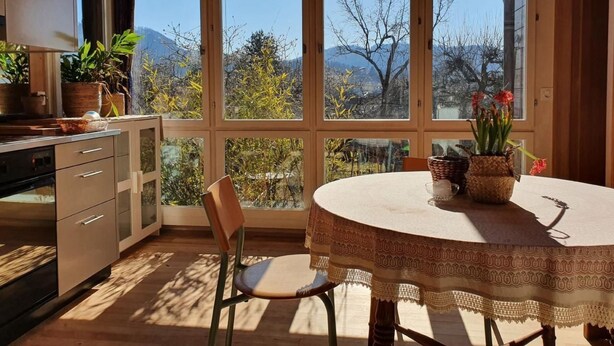 A kitchen with a dining table and chairs, large windows with a view of trees and mountains, and potted plants on the windowsill.