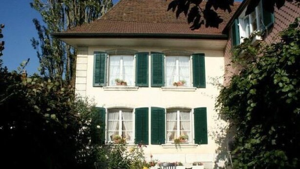 Ein zweistöckiges Haus mit grünen Fensterläden, einer Veranda und einem kleinen Garten mit Tisch und Stühlen.