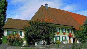 Ein zweist öckiges Gebäude mit geneigtem Dach, grünen Fensterläden und einem kleinen Vorbereich mit roten Blumen.